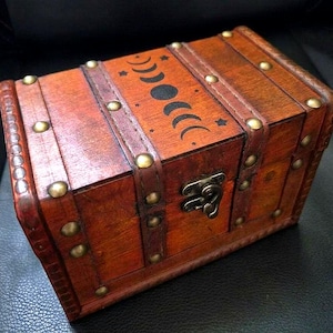 May include: A rectangular wooden chest with a dark brown finish, featuring a hinged lid decorated with a black moon phase graphic and small stars. The box is reinforced with brown leather-like straps secured by round brass-toned metal studs.