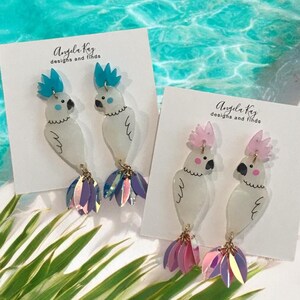 Könnte beinhalten: Ein Paar weiße Kakadu-Ohrringe mit schillernden Federn. Die Ohrringe befinden sich auf einer weißen Karte mit dem Text "Angela Kay designs and finds".