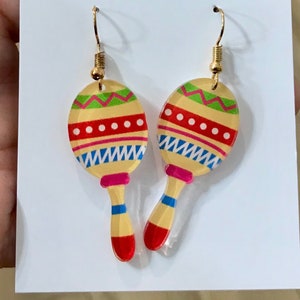 May include: A pair of gold maraca earrings with a colorful design. The maracas are yellow, red, green, blue, and white. The earrings are on a white card with the text "Angela Kay designs and finds".