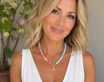 May include: A beaded necklace featuring a gradient of pink to light blue beads with a gold-toned circular clasp and a central gold-toned letter A pendant. The necklace is worn around the neck, showcasing the transition from warm pink to cool blue.