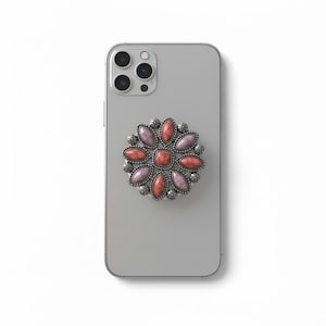 May include: A silver smartphone with a decorative phone grip attached. The grip is a flower design with a silver border and features red and lavender-colored stones. The phone has a triple-lens camera.
