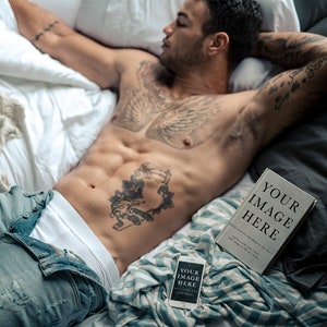 May include: A shirtless man with tattoos lies on a bed with a white and blue striped blanket. He is wearing blue jeans and white underwear. A book with the text "YOUR IMAGE HERE" and a phone with the text "YOUR IMAGE HERE" are on the bed.