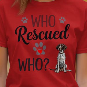 May include: Red t-shirt with the text "WHO Rescued WHO?" in black script and a dog graphic. The design includes paw prints and a heart. The dog is a German Shorthaired Pointer.