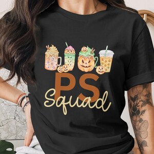 May include: Black t-shirt featuring a watercolor design of four fall-themed beverages, including a pumpkin spice latte and iced coffee. The text "PS Squad" is printed in a mix of brown and yellow script. A festive and fun design.