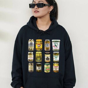 May include: A black hoodie with a graphic print of various pickle jars. The jars have different labels and brands. The hoodie has a drawstring hood and a kangaroo pocket.