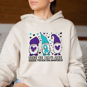 May include: A cream-colored hoodie with a graphic of three gnomes holding purple hearts and a blue ribbon. The text "Gnome One Fights Alone Suicide Prevention Awareness" is printed below the gnomes.