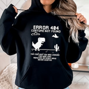 May include: Black hooded sweatshirt with the text "ERROR 404 COSTUME NOT FOUND" and a pixelated dinosaur graphic. The text below reads "THE COSTUME YOU ARE LOOKING FOR WAS LOST REMOVED OR MIGHT HAVE NEVER EXISTED".