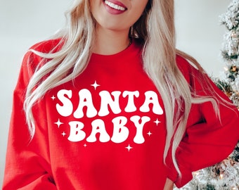May include: A red sweatshirt with the text "SANTA BABY" in white, retro-style lettering. The design includes small white star accents. The sweatshirt is a solid red color.