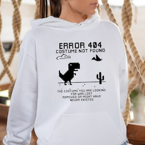 May include: White hoodie with a black graphic print of a dinosaur and the text "ERROR 404 COSTUME NOT FOUND". The text below the dinosaur says "THE COSTUME YOU ARE LOOKING FOR WAS LOST REMOVED OR MIGHT HAVE NEVER EXISTED".