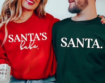 May include: A red sweatshirt with the text "SANTA'S babe." and a green sweatshirt with the text "SANTA." Both sweatshirts are white. The sweatshirts are part of a matching set, perfect for the holiday season.
