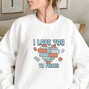 May include: White sweatshirt with a graphic of a heart made of colorful building blocks. The text "I love you to pieces" is printed below the heart.