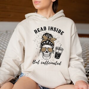 May include: A beige hoodie with a graphic of a skull wearing sunglasses and a leopard print headband. The text "DEAD INSIDE But caffeinated" is printed above and below the skull graphic. The hoodie has a drawstring hood.