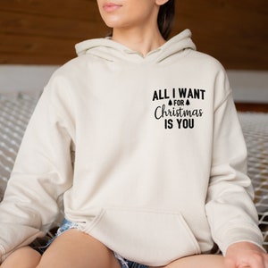 May include: A cream-colored hoodie with a front pocket and drawstring hood. The text "ALL I WANT FOR Christmas IS YOU" is printed in black on the upper chest, with a small Christmas tree graphic above the word "Christmas".