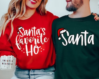 May include: Red and green Christmas sweaters with white text. The red sweater says "Santa's favorite HO" and the green sweater says "Santa". Both sweaters have a Santa hat graphic. The sweaters are worn by a couple.
