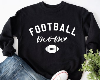 May include: Black sweatshirt with the words "FOOTBALL mom" in white, along with a white football graphic. The sweatshirt is paired with blue jeans and white sneakers, suggesting a casual, sporty style.