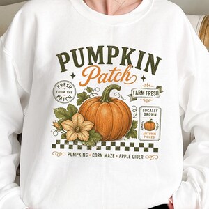 May include: A white crewneck sweatshirt featuring a graphic print with the text PUMPKIN Patch, Fresh from the Patch, Farm Fresh, and Locally Grown Autumn Picked. The design includes a large orange pumpkin, a yellow flower, and a checkered border.