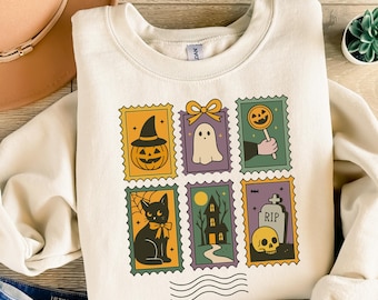 May include: Cream-colored sweatshirt featuring six Halloween-themed postage stamp designs. The stamps depict a jack-o'-lantern, ghost, lollipop, black cat, haunted house, and skull with a tombstone. The sweatshirt is folded on a wooden surface.