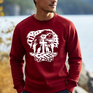 May include: Red sweatshirt with a white graphic design featuring an eagle, a cross, and three soldiers standing in front of an American flag.