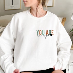 Może przedstawiać: Biała bluza z napisem "YOU ARE enough" w kolorowych literach z małym czarnym sercem.