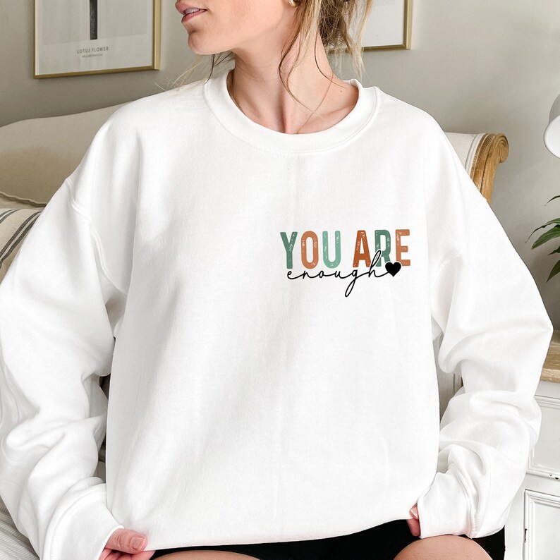 Może przedstawiać: Biała bluza z napisem "YOU ARE enough" w kolorowych literach z małym czarnym sercem.