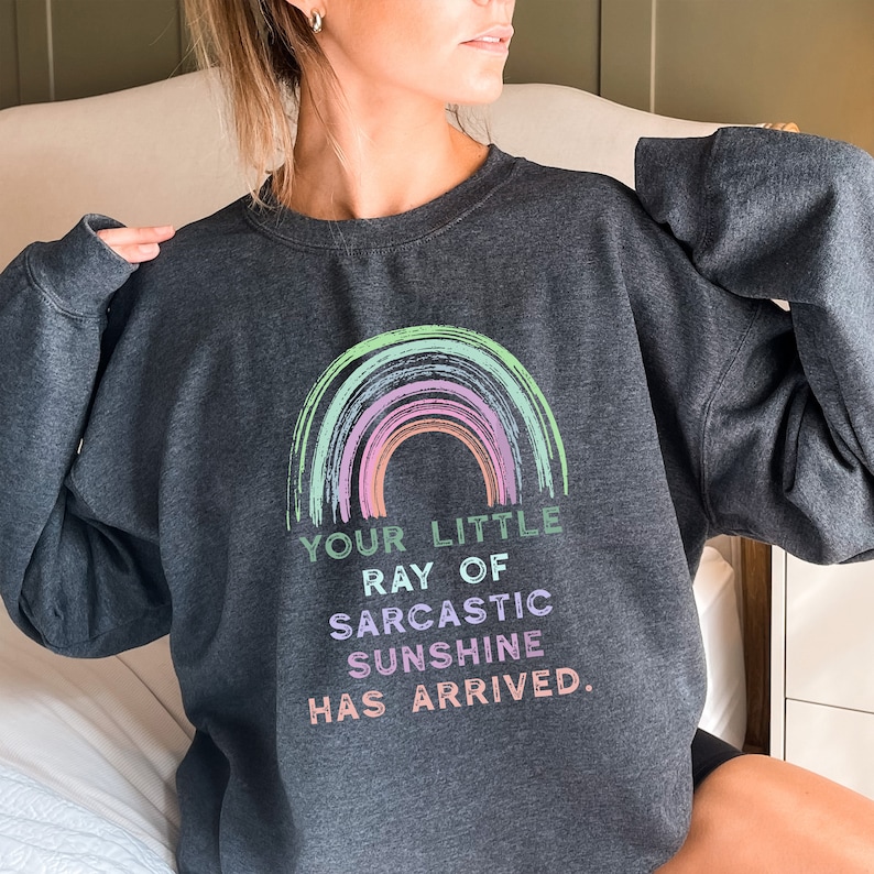 May include: A dark grey sweatshirt with a rainbow design and the text "Your little ray of sarcastic sunshine has arrived."