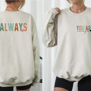 Może przedstawiać: Kremowa bluza z napisem "ALWAYS" w kolorowych literach z tyłu i r&oacute;żowym sercem. Prz&oacute;d bluzy ma napis "YOU ARE enough" w kolorze czarnym z małym czarnym sercem.