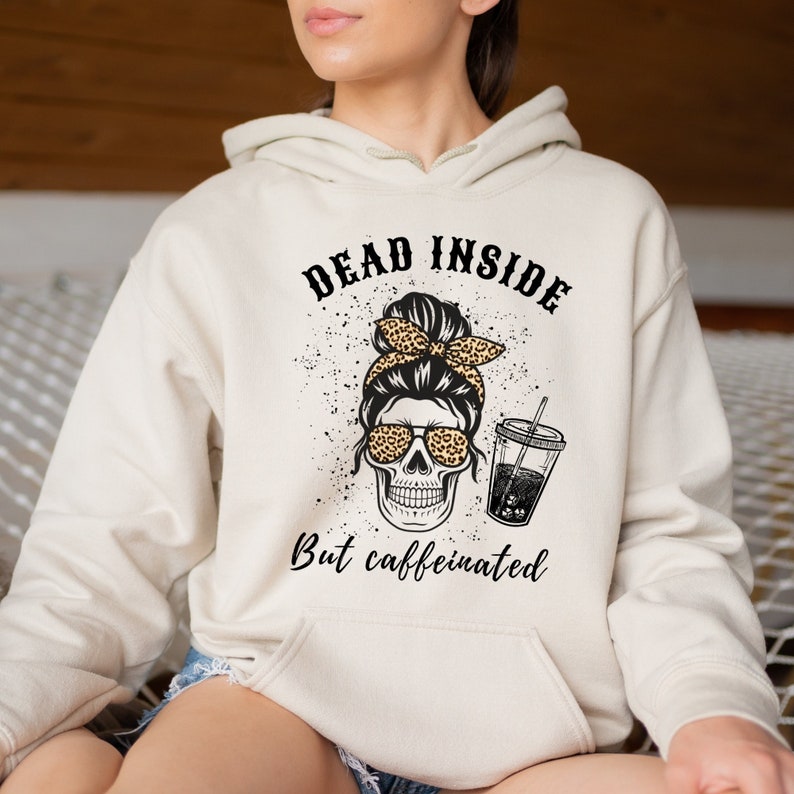 May include: A beige hoodie with a graphic of a skull wearing sunglasses and a leopard print headband. The text "DEAD INSIDE But caffeinated" is printed above and below the skull graphic. The hoodie has a drawstring hood.