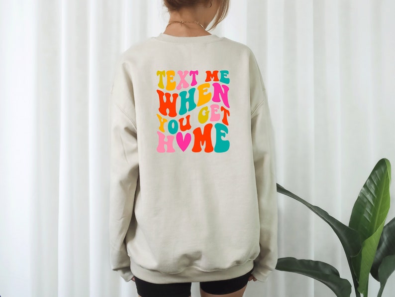 May include: A beige sweatshirt with a colourful graphic on the back that says "Text Me When You Get Home" with a pink heart.