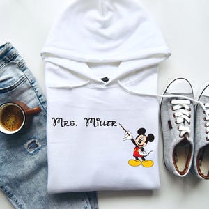 Peut inclure: Sweat-shirt &agrave; capuche blanc avec un motif de Mickey Mouse et le texte "Mrs. Miller" imprim&eacute; sur le devant.
