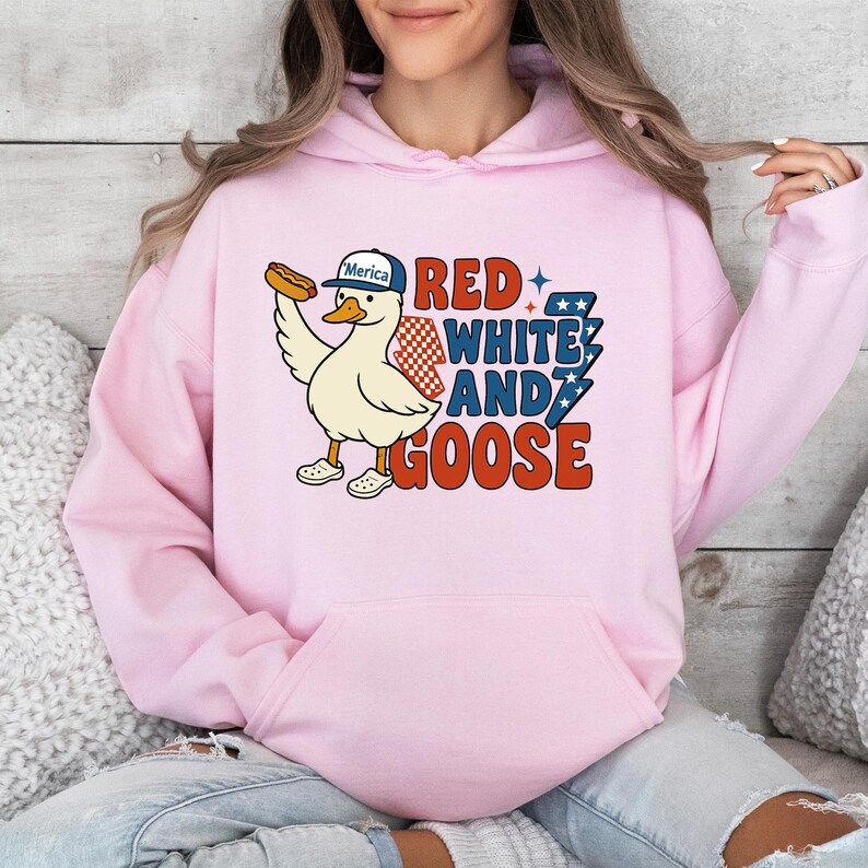 May include: A light pink hoodie featuring a cartoon goose wearing a baseball cap that says "Merica" and holding a hot dog. The text "RED WHITE AND GOOSE" is printed in red, white, and blue.