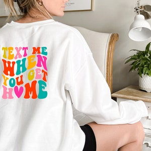 May include: White sweatshirt with a colourful retro-style graphic that says "Text Me When You Get Home" with a pink heart.