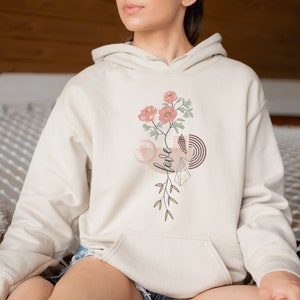 May include: A cream-colored hoodie with a floral design and the word "love" in a stylized font. The design features pink flowers, green leaves, and geometric shapes in brown and gold.