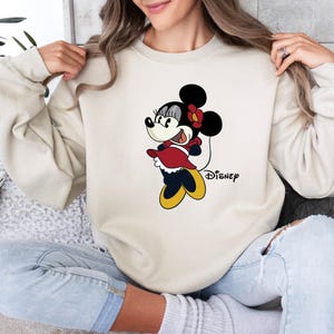 May include: A cream-colored sweatshirt with a cartoon image of Minnie Mouse in a red dress and a black bow. The text "Disney" is printed below the image.