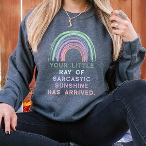 May include: A dark gray sweatshirt with a rainbow graphic and the text "Your little ray of sarcastic sunshine has arrived."