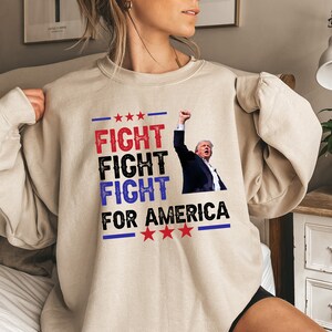 May include: A beige sweatshirt with a graphic print of a person with their fist raised in the air. The text on the sweatshirt reads "Fight Fight Fight For America" with stars above and below the text.