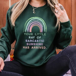 May include: A dark green sweatshirt with a rainbow graphic and the text "Your little ray of sarcastic sunshine has arrived."