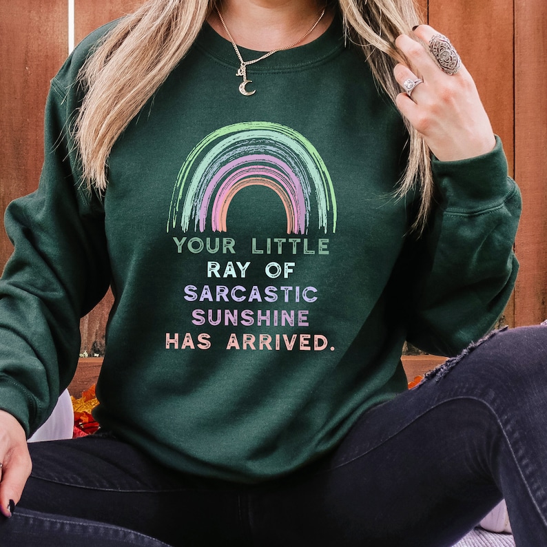 May include: A dark green sweatshirt with a rainbow graphic and the text "Your little ray of sarcastic sunshine has arrived."