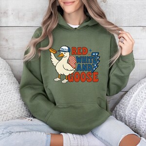 May include: Green hooded sweatshirt with a graphic of a cartoon goose wearing a hat that says "Merica" and holding a hot dog. The text "RED WHITE AND GOOSE" is printed on the front in red, white, and blue.