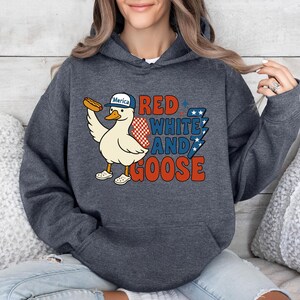 May include: Dark gray hoodie featuring a cartoon goose wearing a baseball cap that says "Merica" and holding a hot dog. The text "Red, White, and Goose" is printed in red, white, and blue. The hoodie is a comfortable, casual style.