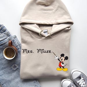 Peut inclure: Un sweat &agrave; capuche beige avec un motif Mickey Mouse noir et le texte "MRS. MILLER" imprim&eacute; sur le devant.