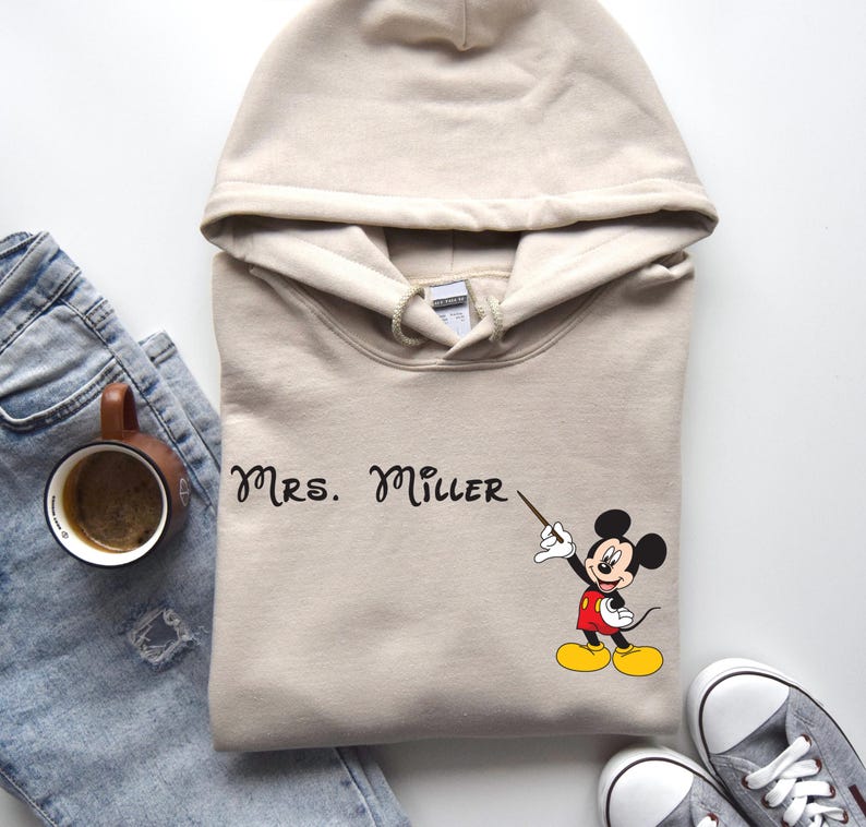 Peut inclure: Un sweat &agrave; capuche beige avec un motif Mickey Mouse noir et le texte "MRS. MILLER" imprim&eacute; sur le devant.