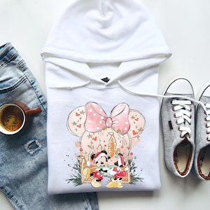 Peut inclure: Sweat &agrave; capuche blanc avec un motif Minnie Mouse, orn&eacute; d'un n&oelig;ud rose et d'un ch&acirc;teau. Le graphisme comprend Mickey et Minnie Mouse en tenues de f&ecirc;te. Des baskets grises et une tasse de caf&eacute; sont &eacute;galement visibles.
