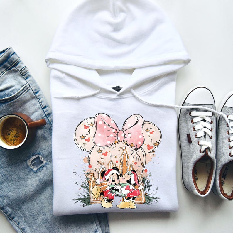 Peut inclure: Sweat &agrave; capuche blanc avec un motif Minnie Mouse, orn&eacute; d'un n&oelig;ud rose et d'un ch&acirc;teau. Le graphisme comprend Mickey et Minnie Mouse en tenues de f&ecirc;te. Des baskets grises et une tasse de caf&eacute; sont &eacute;galement visibles.