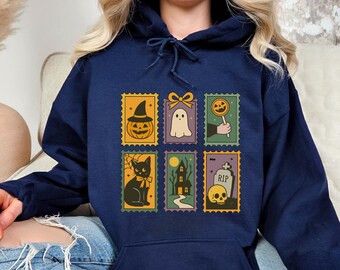 May include: Navy blue hooded sweatshirt featuring six Halloween-themed postage stamp designs. The stamps depict a jack-o'-lantern, ghost, lollipop, black cat, haunted house, and skull with a tombstone. The sweatshirt has a drawstring hood and a kangaroo pocket.