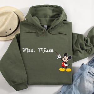 Peut inclure: Un sweat-shirt &agrave; capuche vert avec le texte "Mrs. Miller" et une image de dessin anim&eacute; de Mickey Mouse tenant une baguette de chef d'orchestre.