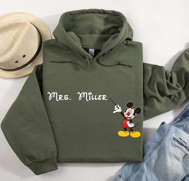 Peut inclure: Un sweat-shirt &agrave; capuche vert avec le texte "Mrs. Miller" et une image de dessin anim&eacute; de Mickey Mouse tenant une baguette de chef d'orchestre.