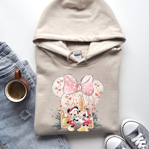 Peut inclure: Sweat &agrave; capuche beige avec un motif Minnie Mouse. Le motif comprend un n&oelig;ud rose &agrave; pois, des accents sur le th&egrave;me de No&euml;l et Mickey et Minnie Mouse en tenues de f&ecirc;te, avec un ch&acirc;teau et des feux d'artifice.