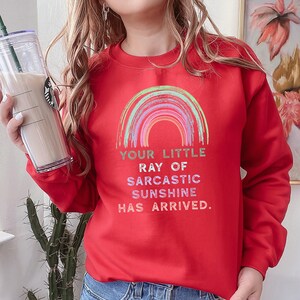 May include: Red sweatshirt with a rainbow graphic and the text "Your little ray of sarcastic sunshine has arrived."