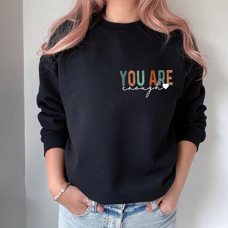 Może przedstawiać: Czarna bluza z napisem "YOU ARE enough" w kolorowych literach z małym białym sercem.