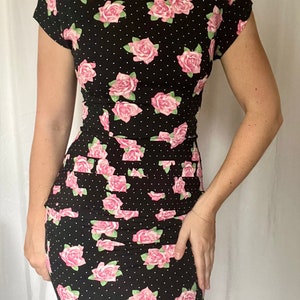 Puede incluir: Un vestido negro con estampado de rosas rosas y lunares blancos. El vestido tiene mangas cortas y una silueta ajustada.